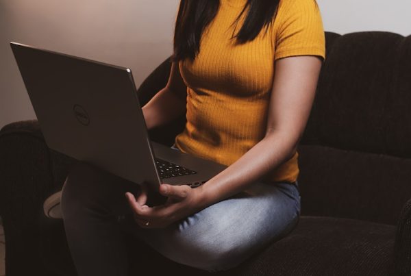 woman in yellow shirt and blue denim jeans sitting on black sofa using macbook