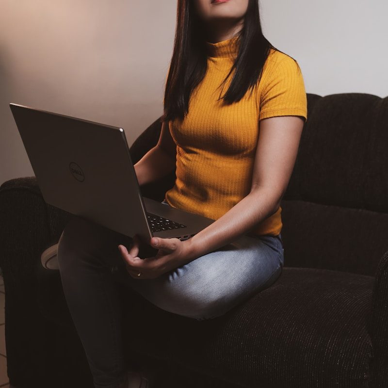 woman in yellow shirt and blue denim jeans sitting on black sofa using macbook